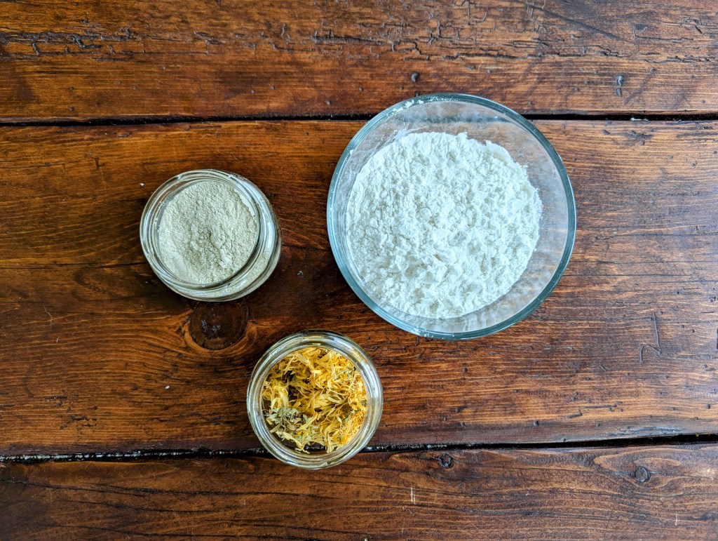 Natural baby powder ingredients including arrowroot powder, bentonite clay, and calendula flowers on a wooden table
